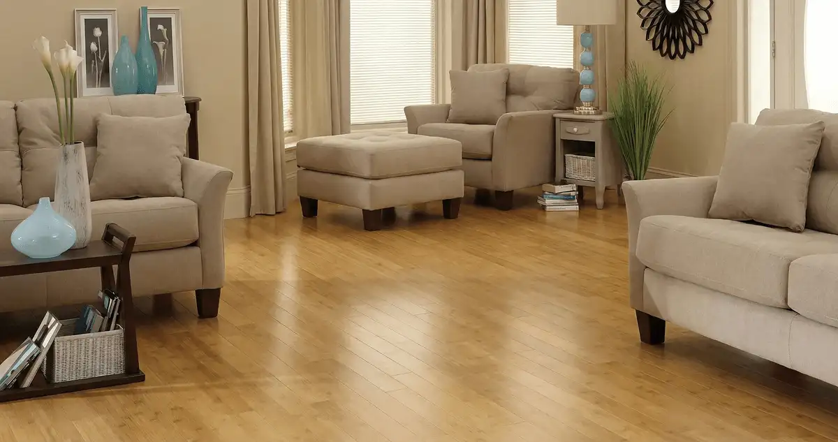 A living room with professionally installed bamboo flooring, beige sofas, a matching ottoman, a side table with a blue lamp, vases, framed pictures, and large windows with blinds and curtains in Albany, NY